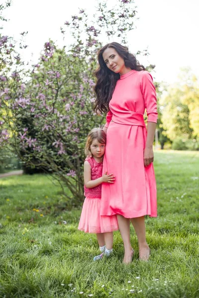 A young beautiful mother with a little daughter is walking along the spring park. Portrait of a mother and a little daughter near the blooming magnolia. Spring. A happy family. Heat. — Stock Photo, Image