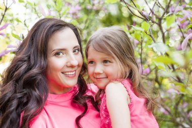 Küçük kızı olan genç güzel bir anne bahar park boyunca yürüyor. Bir anne ve çiçek açan magnolia yakınındaki küçük bir kız portresi. Bahar. Mutlu bir aile. Isı.