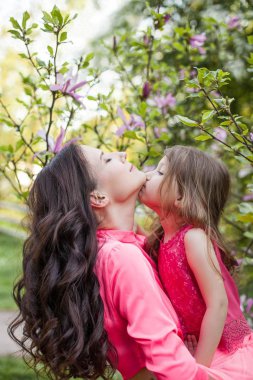 Küçük kızı olan genç güzel bir anne bahar park boyunca yürüyor. Bir anne ve çiçek açan magnolia yakınındaki küçük bir kız portresi. Bahar. Mutlu bir aile. Isı.