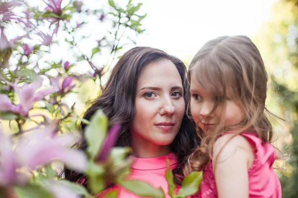 A young beautiful mother with a little daughter is walking along the spring park. Portrait of a mother and a little daughter near the blooming magnolia. Spring. A happy family. Heat. — Stock Photo, Image