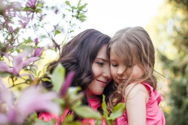 Una joven y hermosa madre con una hija pequeña está caminando por el parque de primavera. Retrato de una madre y una pequeña hija cerca de la floreciente magnolia. Primavera. Una familia feliz. Calor . —  Fotos de Stock