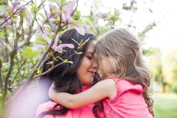 A young beautiful mother with a little daughter is walking along the spring park. Portrait of a mother and a little daughter near the blooming magnolia. Spring. A happy family. Heat. — Stock Photo, Image