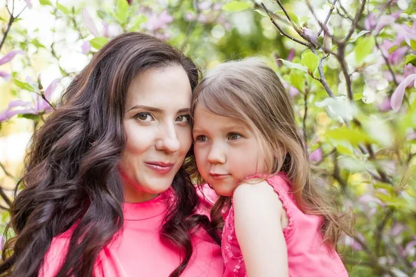 A young beautiful mother with a little daughter is walking along the spring park. Portrait of a mother and a little daughter near the blooming magnolia. Spring. A happy family. Heat. — Stock Photo, Image