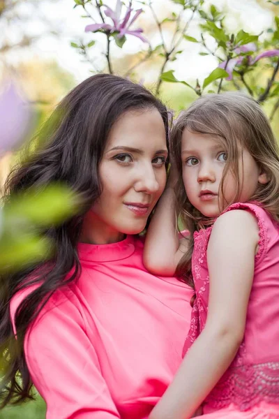 Küçük kızı olan genç güzel bir anne bahar park boyunca yürüyor. Bir anne ve çiçek açan magnolia yakınındaki küçük bir kız portresi. Bahar. Mutlu bir aile. Isı.