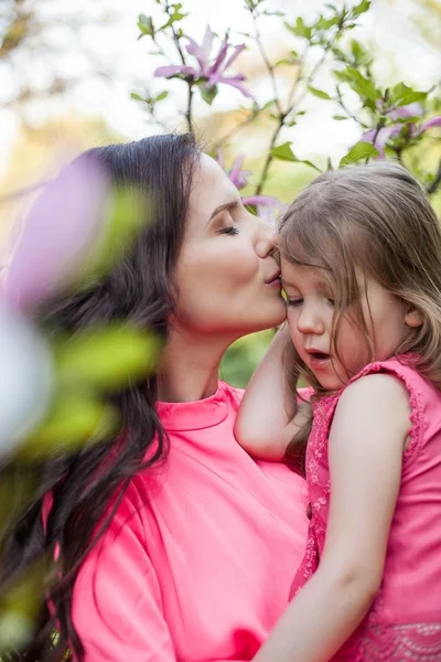 Una joven y hermosa madre con una hija pequeña está caminando por el parque de primavera. Retrato de una madre y una pequeña hija cerca de la floreciente magnolia. Primavera. Una familia feliz. Calor . —  Fotos de Stock