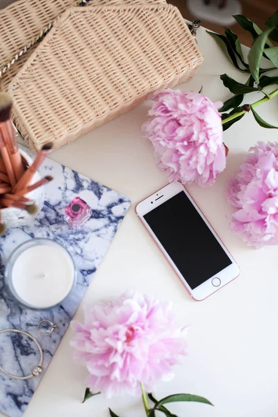Peonies. Bouquet of pink peonies, candle, wicker bag, scarf, perfume, jewelry, phone on white table. Cosmetic table. Spring.