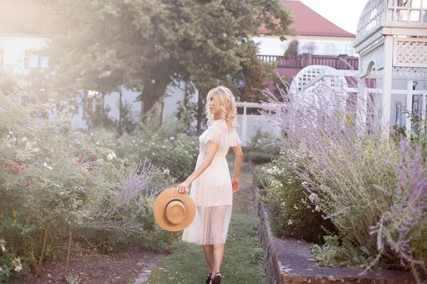 在阳光下的女孩的肖像 年轻的金发女郎正在一个五颜六色的花园里散步 一个年轻的女人拿着花和一顶草帽 — 图库照片