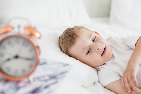 La sveglia è sul comodino vicino al letto. Un bambino è sdraiato su un letto bianco. Svegliati. Buongiorno. Accogliente. Scuola . — Foto Stock