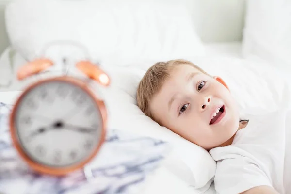 La sveglia è sul comodino vicino al letto. Un bambino è sdraiato su un letto bianco. Svegliati. Buongiorno. Accogliente. Scuola . — Foto Stock