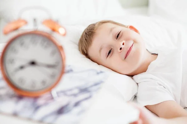 The alarm clock is on the bedside table by the bed. A little boy is lying on a white bed. Wake up. Morning. Cozy. School.