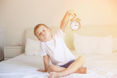 Sevimli küçük bir çocuk yatak odasında yatağın üstüne oturan ve bir çalar saat tutuyor. Güneş ışınları. Sabah. Uyandır. Rahat.