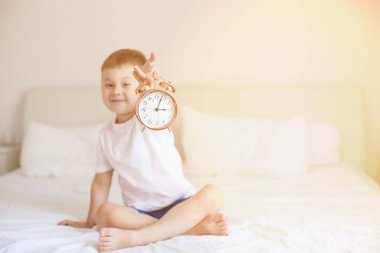 Sevimli küçük bir çocuk yatak odasında yatağın üstüne oturan ve bir çalar saat tutuyor. Güneş ışınları. Sabah. Uyandır. Rahat.