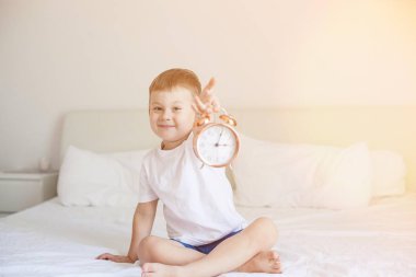 Sevimli küçük bir çocuk yatak odasında yatağın üstüne oturan ve bir çalar saat tutuyor. Güneş ışınları. Sabah. Uyandır. Rahat.