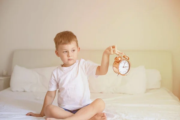 Un niño lindo está sentado en el dormitorio en la cama y sostiene un reloj despertador. Rayos solares. Buenos días. Despierta. Acogedor . — Foto de Stock