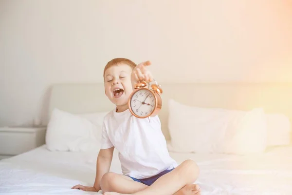 Un bambino carino è seduto nella camera da letto sul letto e tiene una sveglia. I raggi del sole. Buongiorno. Svegliati. Accogliente . — Foto Stock