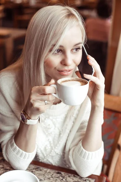 Retrato de una hermosa rubia joven. Una joven feliz en un suéter de punto está sentada en un café bebiendo capuchino y hablando por teléfono. Otoño. El invierno. Acogedor . — Foto de Stock