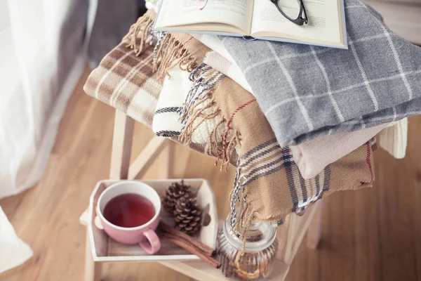 Плайды. Чашку горячего чая. Осенний уютный интерьер. На деревянном стуле - стопка теплых одеял. Свечи, листья, шишки, корзина, корица. Книга и очки. Осень. Зима . — стоковое фото