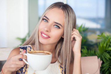 Genç güzel sarışın mavi gözlü bir kafede oturuyor. Genç bir kadın bir kapuçino kupa onun elinde tutar. Mutlu bir kadın portresi. Moda. Makyaj. Yaz.