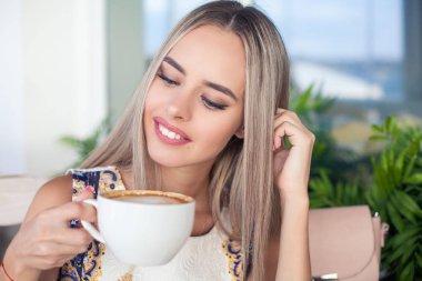 Genç güzel sarışın mavi gözlü bir kafede oturuyor. Genç bir kadın bir kapuçino kupa onun elinde tutar. Mutlu bir kadın portresi. Moda. Makyaj. Yaz.