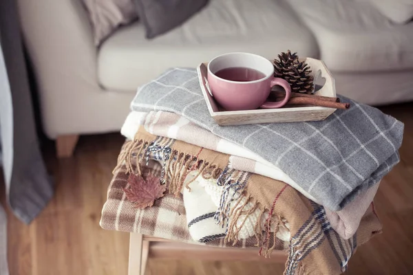 Plaids. A cup of hot tea. Autumn cozy interior. On a wooden chair is a stack of warm blankets. Candles, leaves, cones, basket, cinnamon. Book and glasses. Autumn.