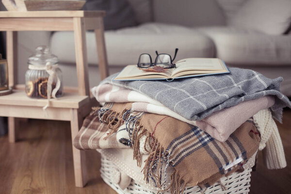 Plaids. A cup of hot tea. Autumn cozy interior. On a wooden chair is a stack of warm blankets. Candles, leaves, cones, basket, cinnamon. Book and glasses. Autumn.