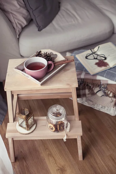 Plaids. A cup of hot tea. Autumn cozy interior. On a wooden chair is a stack of warm blankets. Candles, leaves, cones, basket, cinnamon. Book and glasses. Autumn.