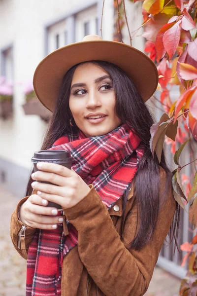 Young beautiful Turkish woman walks in a European city. A girl holds a glass of coffee. Happy brunette in a hat. Autumn. Fashion.