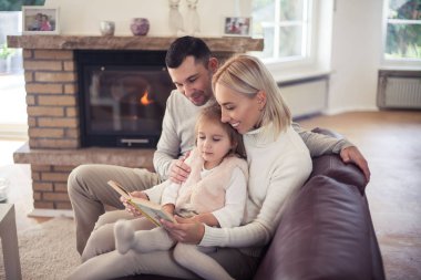 Şöminenin yanında oturan hoş bir aile. Anne, baba ve kız bir kitap okuyor. Annelik. Aile. Sıcak sonbahar. Akşam rahat. Ana sayfa.