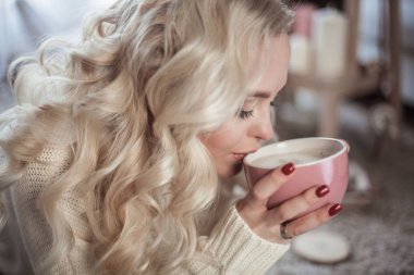 Güzel bir genç kadın rahat ev portresi. Güzel sarışın kız Örgü elbise bir kapuçino içiyor ve bir kitap okuma cam kenarında oturuyor. Sonbahar dekorasyonu. Rahat. Kış. Sonbahar.