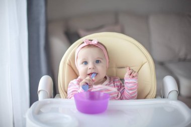 Sevimli küçük kız 7 aylık bir çocuk sandalyede oturur ve bir kaşık tutar. Pembe elbiseli mavi gözlü Bebek portresi. Bebekler için yemek. Rahat.