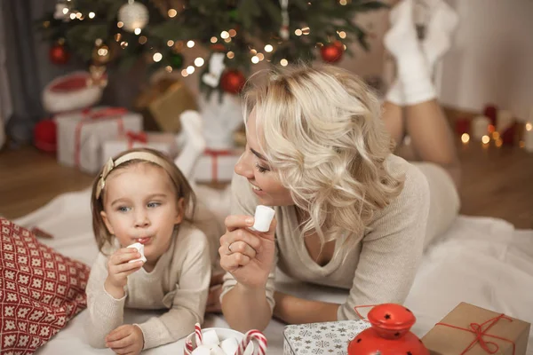 可爱的小女孩和她年轻的母亲躺在圣诞树附近吃糖果 圣诞节 新的一年 — 图库照片