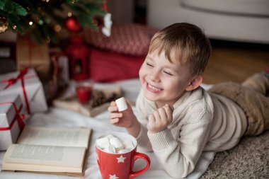 Küçük mutlu sevimli çocuk Noel ağacının yanında yatıyor, bir kitap okur ve sıcak kakao marshmallows ile içecekler. Çocuk. Noel ruh. Tatil. Noel. Yeni yıl. Rahat.