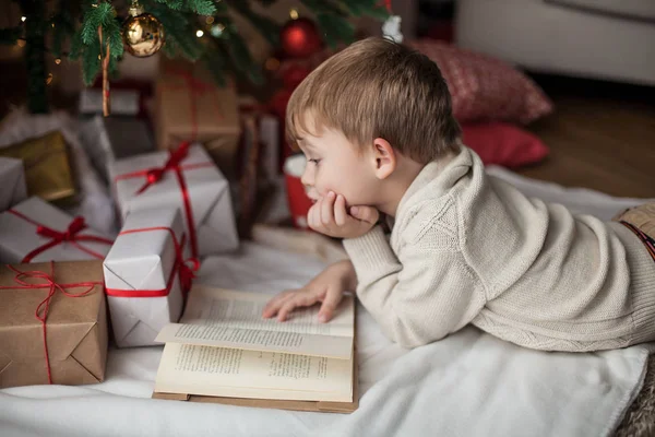 Pequeno menino bonito feliz fica perto da árvore de Natal, lê um livro e bebe cacau quente com marshmallows. Criança. Humor de Natal. Férias. No Natal. Ano Novo. Aconchegante . — Fotografia de Stock