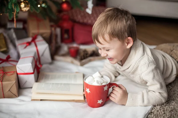 Pequeno menino bonito feliz fica perto da árvore de Natal, lê um livro e bebe cacau quente com marshmallows. Criança. Humor de Natal. Férias. No Natal. Ano Novo. Aconchegante . — Fotografia de Stock