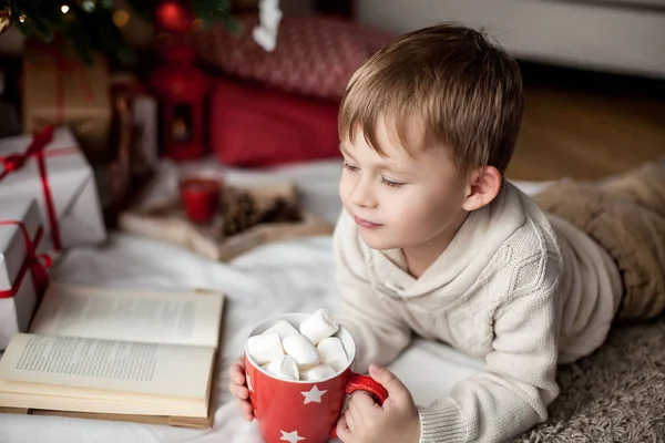 Šťastný roztomilý chlapeček leží poblíž vánoční strom, čte knihu a pije horké kakao s marshmallows. Dítě. Vánoční náladu. Dovolená. Vánoce. Nový rok. Cozy. — Stock fotografie