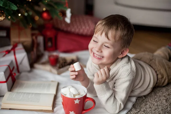 Маленький щасливий милий хлопчик лежить біля ялинки, читає книгу і п'є гаряче какао з зефіром. Дитина. Різдвяний настрій. Відпустка. Різдво. Новий рік. Затишний . — стокове фото