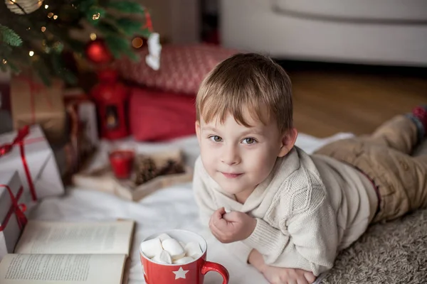 Маленький щасливий милий хлопчик лежить біля ялинки, читає книгу і п'є гаряче какао з зефіром. Дитина. Різдвяний настрій. Відпустка. Різдво. Новий рік. Затишний . — стокове фото