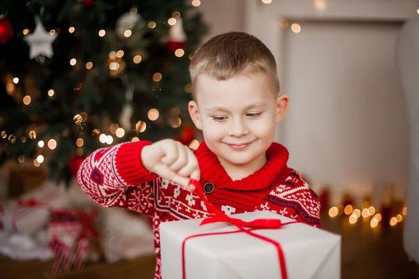 Kırmızı Noel kazağı küçük şirin çocuk Noel ağacının yanında oturur. 4 yıllık bir hediye kutusu ile eski mutlu bir çocuk portresi. Tatil. Noel. Yeni yıl.