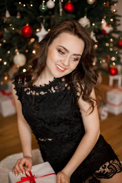 Jovem morena bonita em um vestido preto perto da árvore de Natal. menina feliz abre presentes. Férias. No Natal. Ano Novo . — Fotografia de Stock