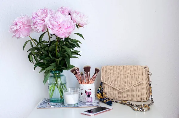 Peonies. Bouquet of pink peonies, candle, wicker bag, scarf, perfume, jewelry, phone on white table Cosmetic table
