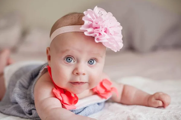 Linda niña de ojos azules en un vestido con una flor en la cabeza. El bebé yace en la cama. Retrato de un niño de 4 meses — Foto de Stock