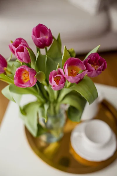 Frühjahrsinnenraum. Ein Strauß Tulpen in einer Glasvase, ein goldenes Tablett, eine Tasse, eine Teekanne, ein rosa Plaid, ein Sofa. gemütlich. — Stockfoto