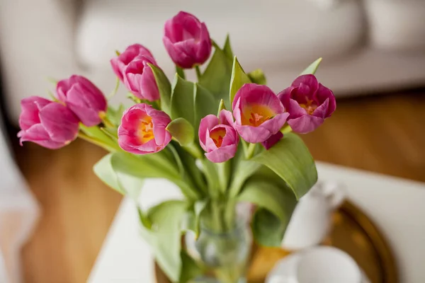 Frühjahrsinnenraum. Ein Strauß Tulpen in einer Glasvase, ein goldenes Tablett, eine Tasse, eine Teekanne, ein rosa Plaid, ein Sofa. gemütlich. — Stockfoto