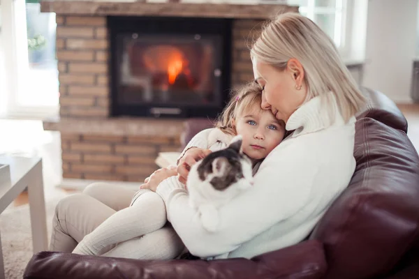 Mooie moeder, dochtertje en een kat zitten op de Bank bij de open haard. Familie in het interieur. Gezellige. — Stockfoto