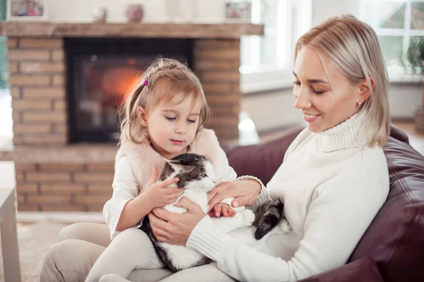 Mãe bonita, filha pequena e um gato estão sentados no sofá perto da lareira. Família no interior da casa. Aconchegante . — Fotografia de Stock