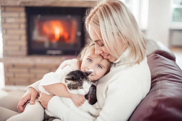 Mooie moeder, dochtertje en een kat zitten op de Bank bij de open haard. Familie in het interieur. Gezellige. — Stockfoto