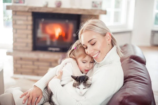 Beautiful mother, little daughter and a cat are sitting on the sofa near the fireplace. Family in the home interior. Cozy. — Stock Photo, Image
