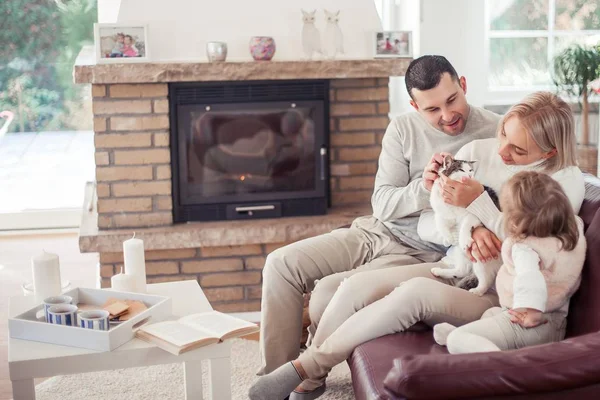 De familie zit op de Bank bij de open haard. Moeder, vader, dochter en kat in het interieur. Gezellige. — Stockfoto