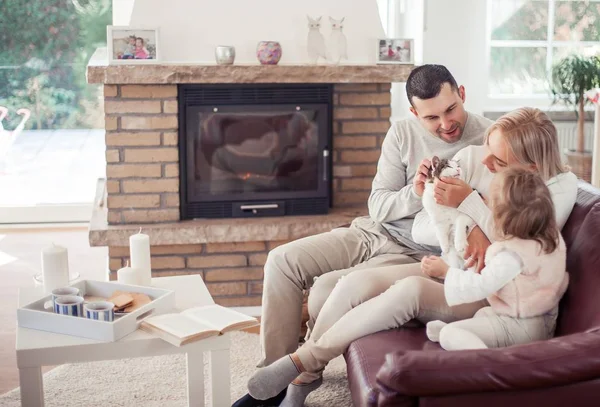 De familie zit op de Bank bij de open haard. Moeder, vader, dochter en kat in het interieur. Gezellige. — Stockfoto