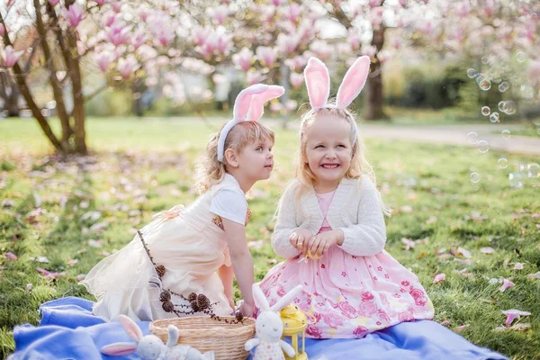 Little cute girls are sitting on the grass near the mogolia. Girls in costumes Easter bunnies. — Stock Photo, Image